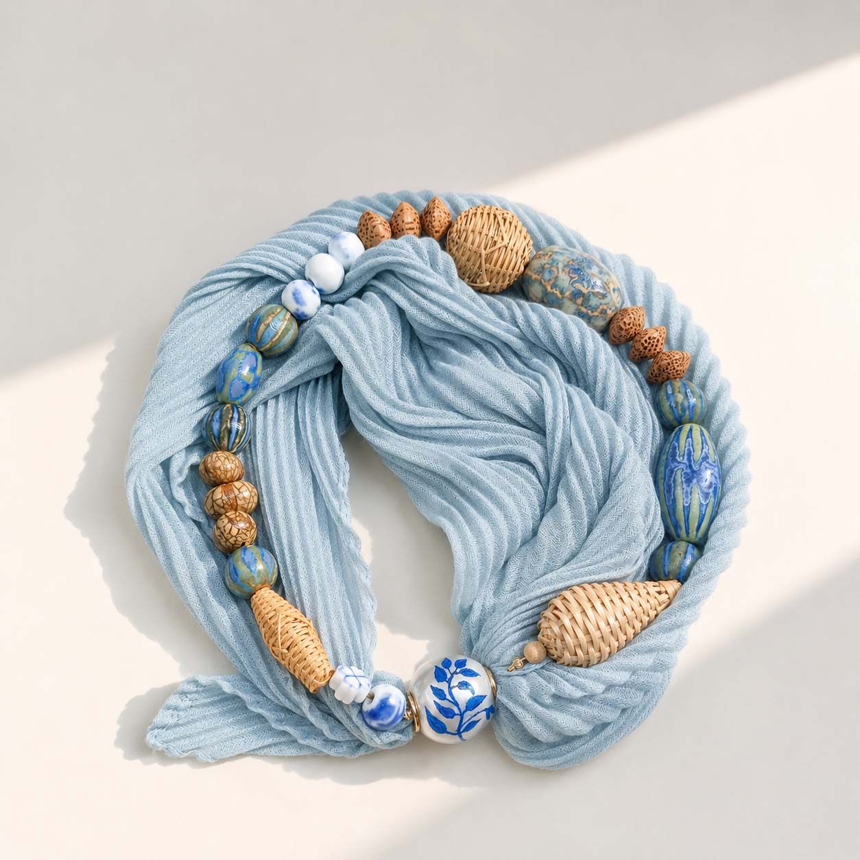 Artisan pleated scarf necklace with navy printed scarf, porcelain beads, woven rattan beads and pearl magnetic clasp on a neutral background.