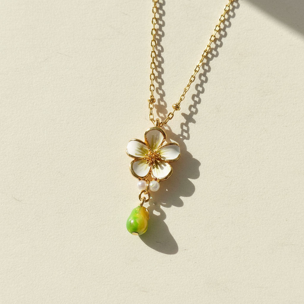Gold necklace with a white flower pendant and green teardrop charm on a beige background
