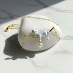 Gold necklace featuring a hand-painted white cat pendant lounging on a bar with a dangling pearl charm.
