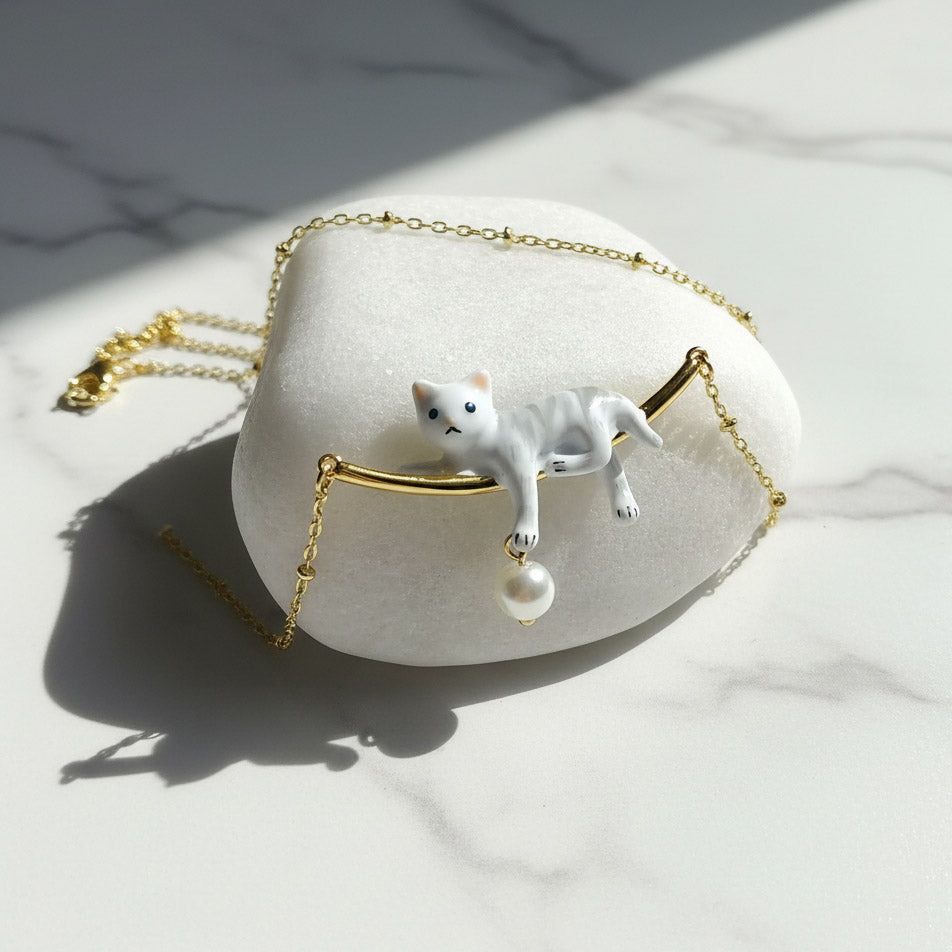 Gold necklace featuring a hand-painted white cat pendant lounging on a bar with a dangling pearl charm.