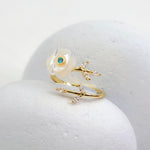 Elegant adjustable gold-plated ring with hand-carved mother-of-pearl flower and blue birthstone accent, displayed on white stone background.
