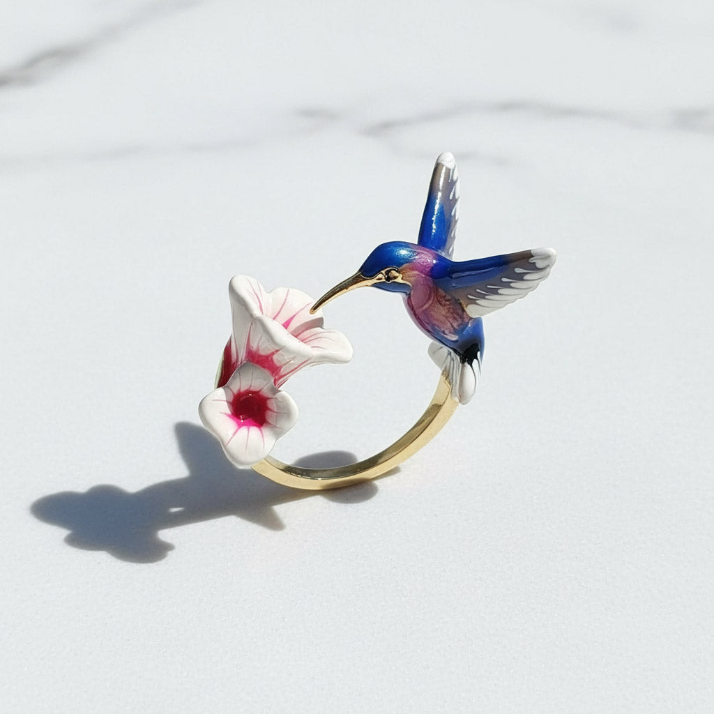 Gold ring with a blue hummingbird and pink flower charm on a light gray background