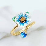 Gold-plated adjustable ring featuring a hand-painted blue and white enamel flower with a matching bud and green leaf detail on white marble background.