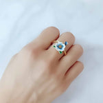 Hand wearing an adjustable gold-plated ring featuring a hand-painted blue and white enamel flower and bud, styled against a soft white background.