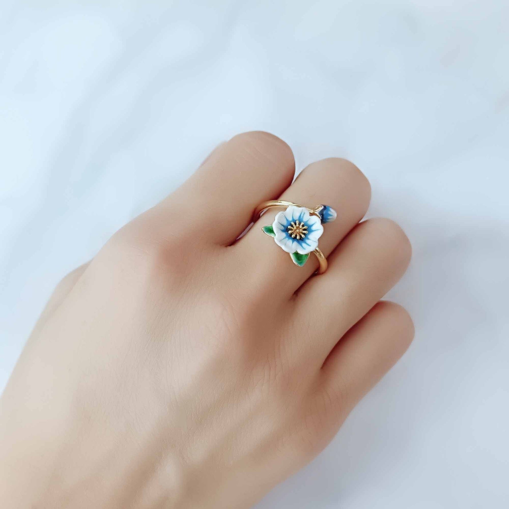 Hand wearing an adjustable gold-plated ring featuring a hand-painted blue and white enamel flower and bud, styled against a soft white background.