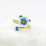 Adjustable gold-plated ring with a hand-painted blue and white enamel flower and bud, styled on a smooth white stone background.