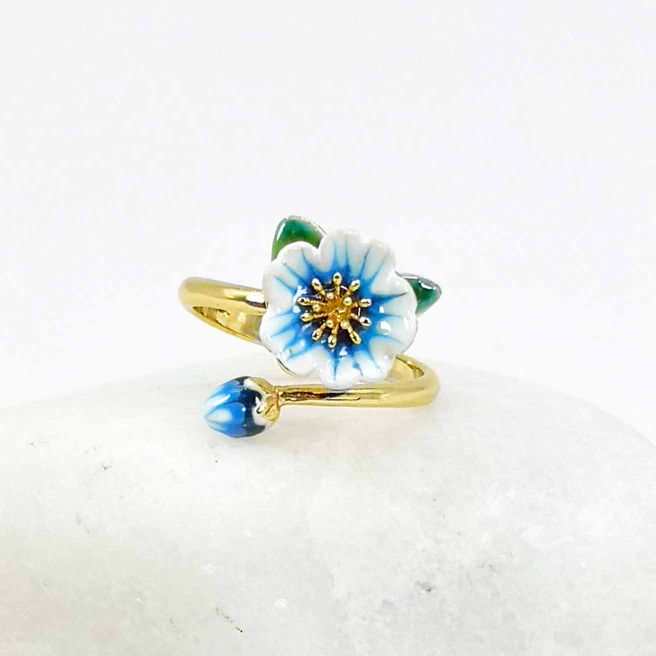 Adjustable gold-plated ring with a hand-painted blue and white enamel flower and bud, styled on a smooth white stone background.