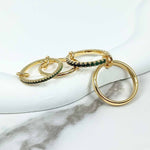 A set of four linked gold-plated rings displayed on a white marble surface. The rings feature green cubic zirconia accents and polished gold finishes, connected by delicate gold links.