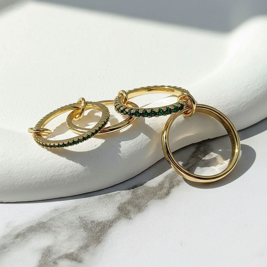 Gold linked ring set with green cubic zirconia stones, three interlocking bands displayed on marble background.