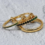 A close-up shot of four linked gold-plated rings on a grey stone background, showcasing green and black cubic zirconia accents with polished and pavé textures.
