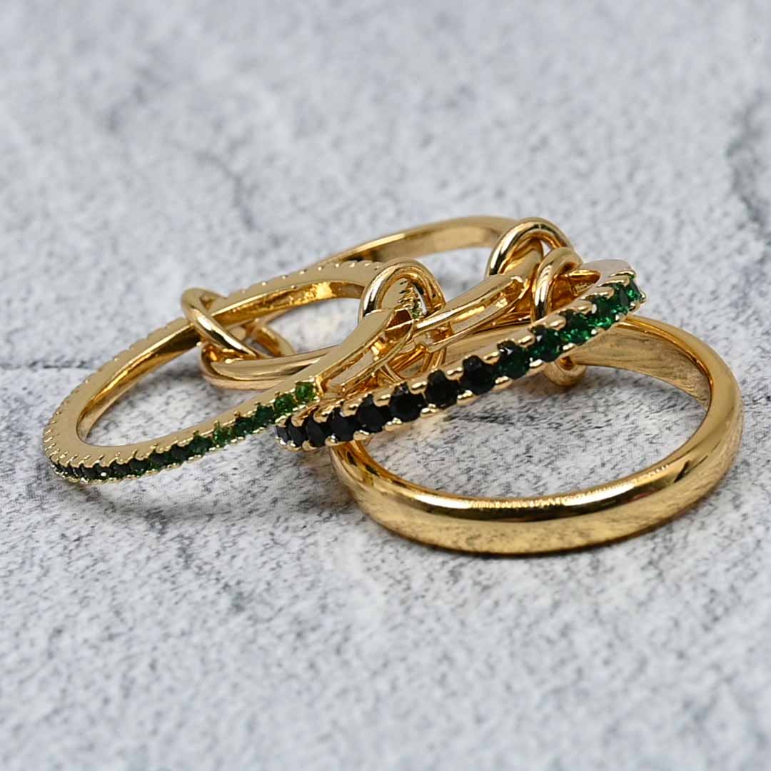 A close-up shot of four linked gold-plated rings on a grey stone background, showcasing green and black cubic zirconia accents with polished and pavé textures.