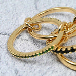 Side view of the linked gold-plated ring set on a grey stone surface, highlighting the emerald cubic zirconia sparkle and the polished gold bands.