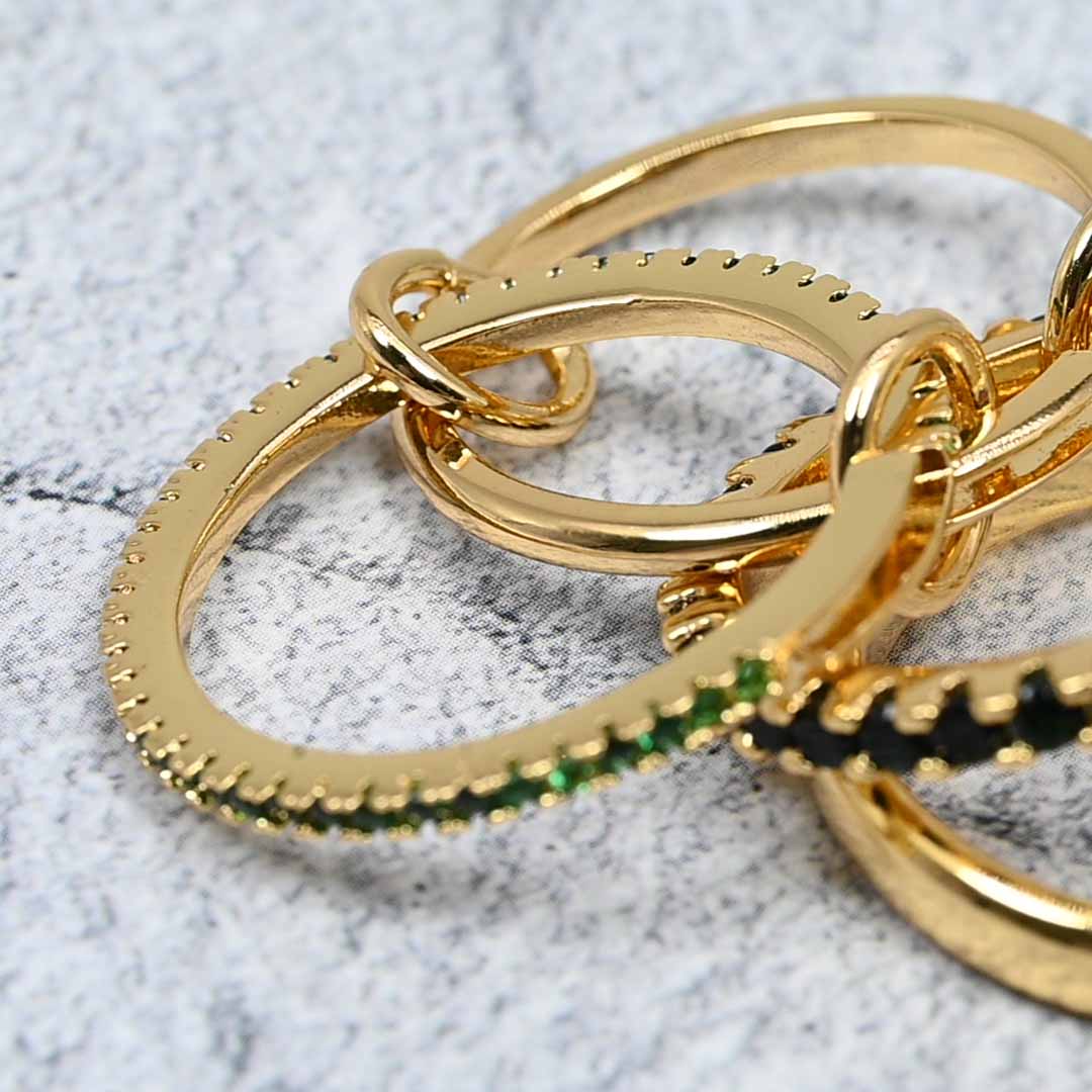 Side view of the linked gold-plated ring set on a grey stone surface, highlighting the emerald cubic zirconia sparkle and the polished gold bands.