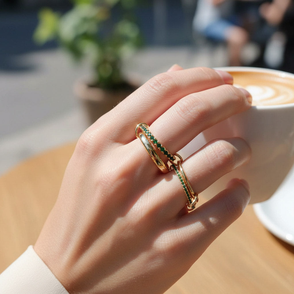 Hand wearing a gold ring with green gemstones in front of a cup of coffee.