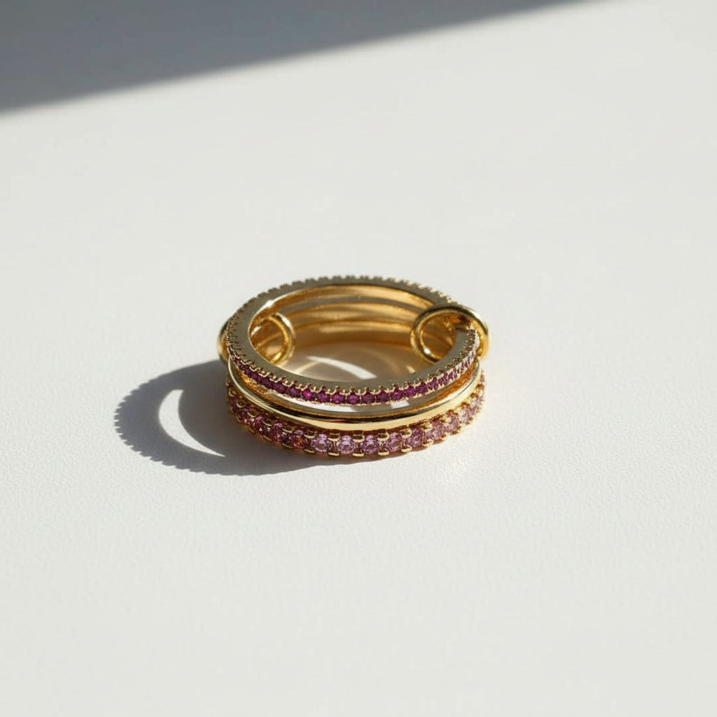 Stacked gold linked rings with pink cubic zirconia stones, featuring three interlocking bands in sunlight.