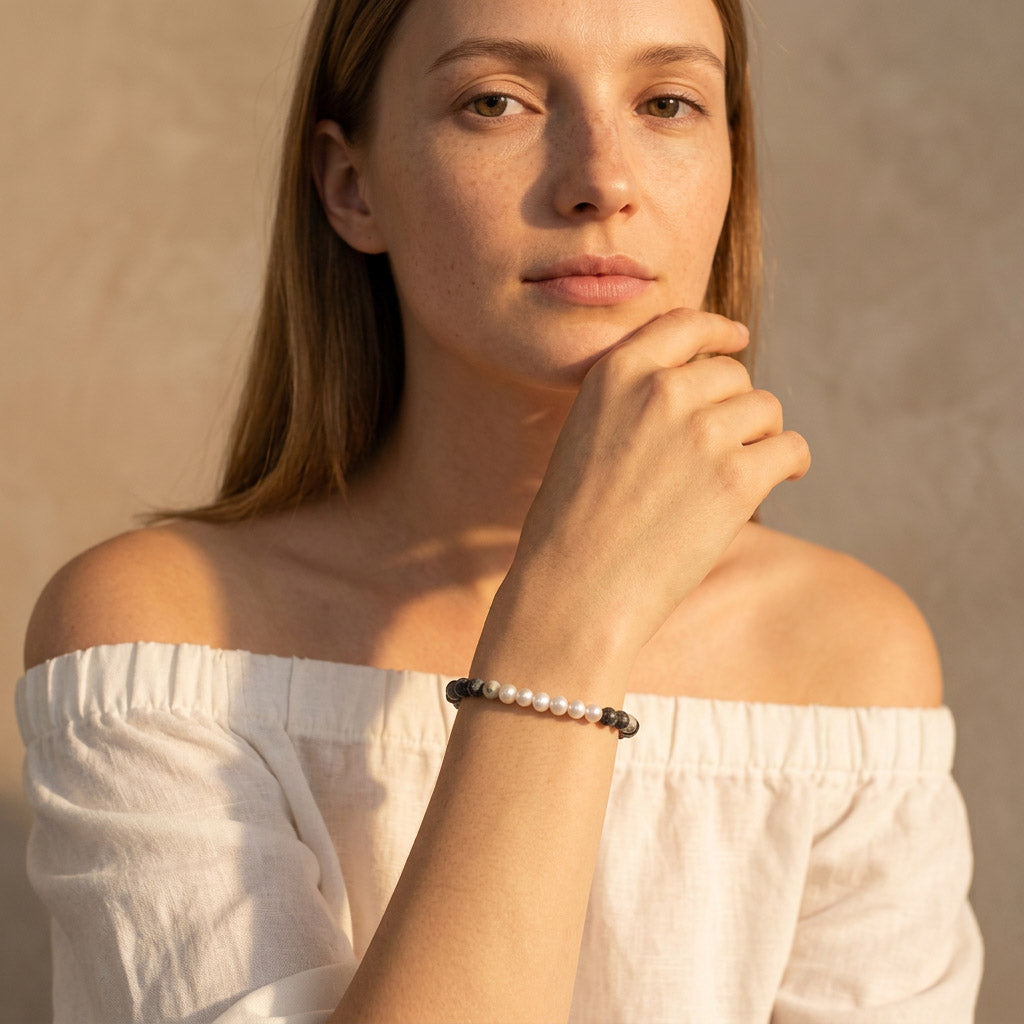 Woman wearing a white off-shoulder top and pearl and zebra gemstone bracelet against a beige background