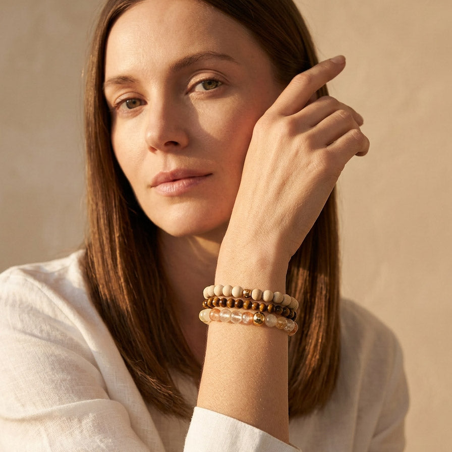 Woman wearing stacked gemstone bracelets with Tiger Eye Rutilated Quartz and camphor wood beads against a beige background
