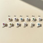 Set of twelve floral earrings with colored gemstones, each labeled for a month on a beige background.
