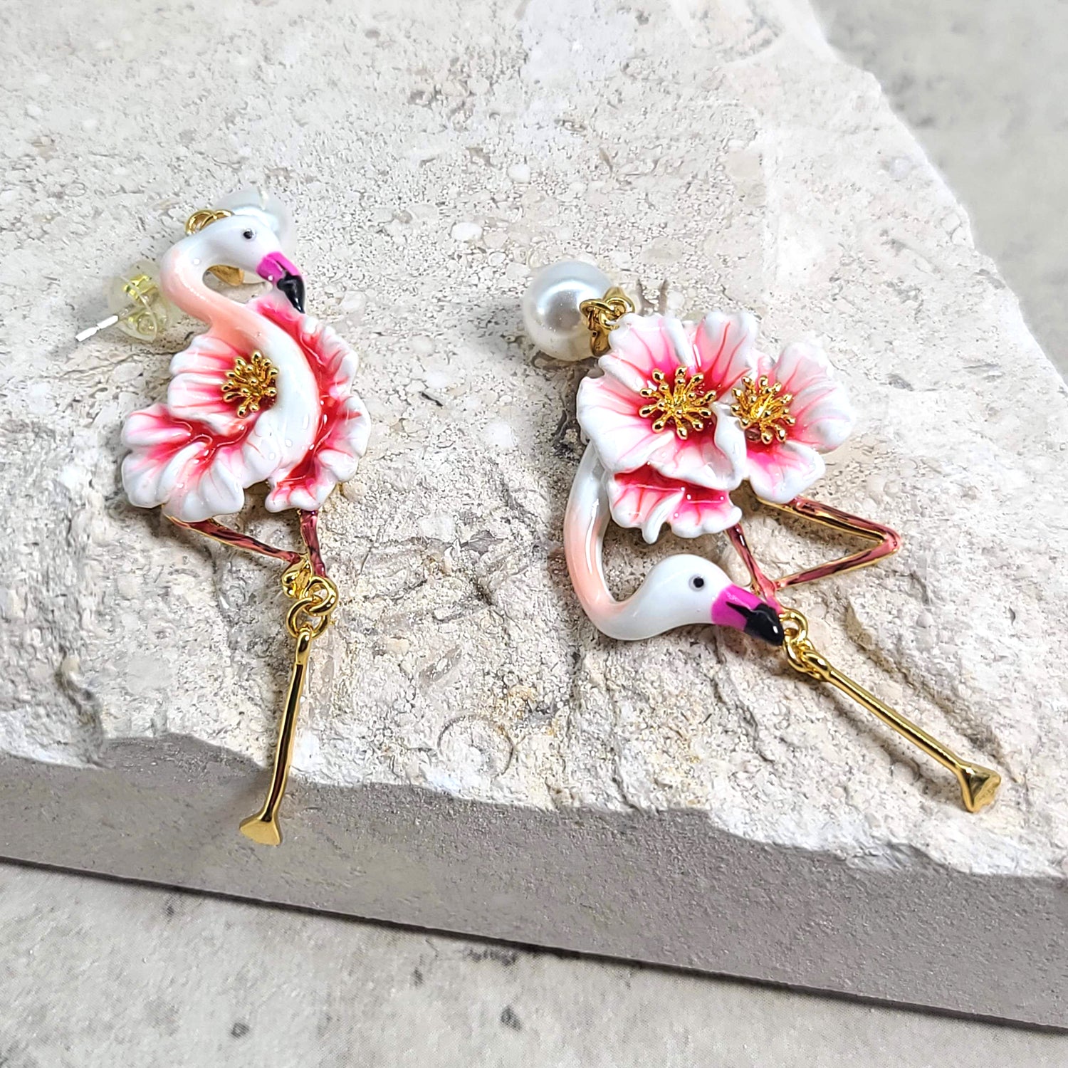 Handcrafted flamingo earrings with enamel pink flowers, pearl stud tops, and gold-tone legs, displayed on a textured beige stone background.