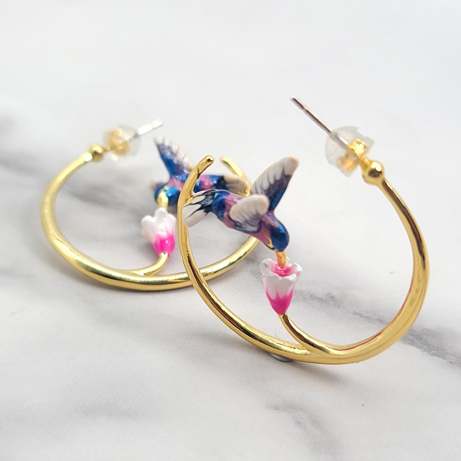  A pair of hoop earrings featuring a delicate enamel-painted hummingbird in mid-flight, feeding from a flower. The earrings are crafted from brass with a silver finish and measure 3.2cm (1.25”) in diameter.